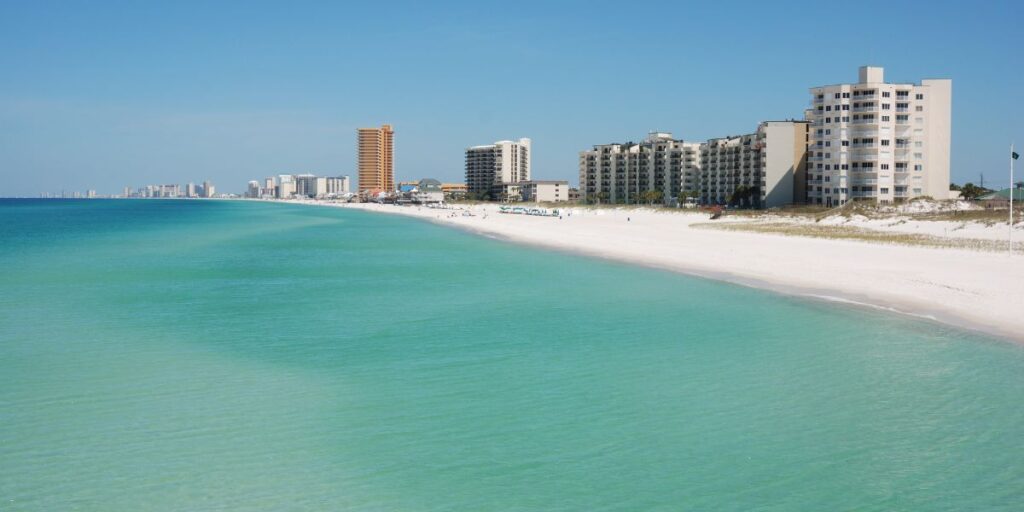 Panama City Beach shoreline with turquoise waters, white sandy beach, and a skyline of beachfront condos. A serene coastal view under a clear blue sky.